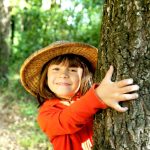 Kind mit Hut umarmt einen Baum.