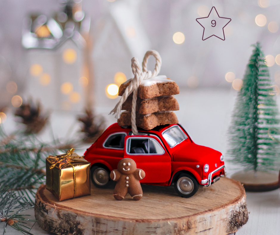 Auf einer Holzscheibe steht ein rotes Auto. Vor dem Auto steht ein links ein golden verpacktes Geschenk und in der Mitte ein kleines Lebkuchenmännchen. Auf dem Dach des Autos sind drei Zimtsterne als Paket geschnürt.