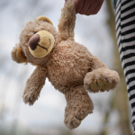 Ein Teddybär baumelt von einer Hand eines Kindes herab, welches einen seiner Arme in der Hand hält.